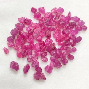 Puede incluir: Un conjunto de gemas de rubí en bruto y sin cortar. Las pequeñas piedras, de forma irregular, muestran un vibrante tono rosa-rojizo. Las gemas están dispersas sobre una superficie blanca, mostrando su estado natural y sin pulir. Estos rubíes son ideales para la creación de joyas.