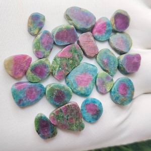May include: A collection of 18 loose, natural, green, blue, and pink gemstones. The stones are irregularly shaped and have a variety of sizes.