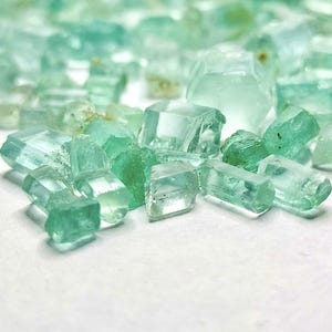 Op de afbeelding: Een verzameling ruwe aquamarijnkristallen. De kristallen variëren in grootte en vorm, met een reeks lichtblauwe en groene tinten. Sommige zijn doorschijnend, andere meer ondoorzichtig. De kristallen zijn verspreid over een witte ondergrond.