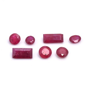 May include: Assortment of seven red ruby gemstones of various shapes and sizes. The gemstones are arranged on a white background.