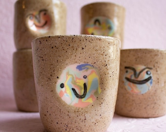 May include: Four ceramic mugs with speckled brown glaze and colorful abstract faces. Each mug has a different face design.