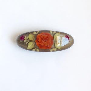 May include: An oval-shaped, brown, resin hair clip with a raised, orange rose and green leaves design.