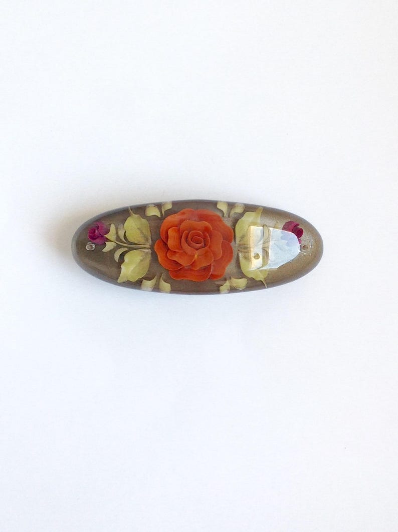May include: An oval-shaped, brown, resin hair clip with a raised, orange rose and green leaves design.