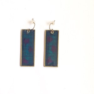 Pode incluir: Um par de brincos retangulares pendentes com acabamento em esmalte azul escuro e roxo. Os brincos têm uma moldura de metal dourado.