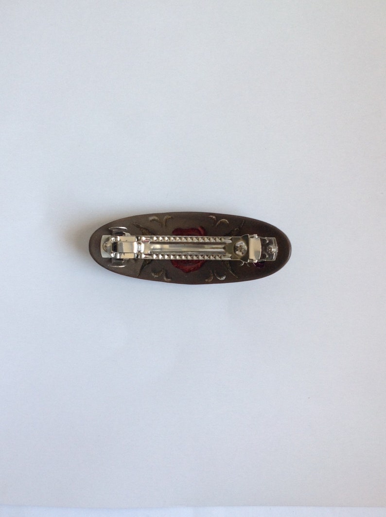 May include: A brown oval hair clip with a silver metal clasp and a red rose design.