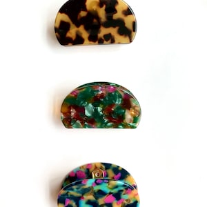 May include: Three half-circle hair clips with different patterned designs. The top clip is brown and black, the middle clip is green and red, and the bottom clip is blue, pink, and black.