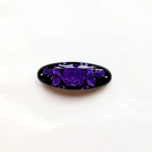 May include: A black oval hair clip with a purple floral design. The design features a large rose with smaller flowers and leaves.