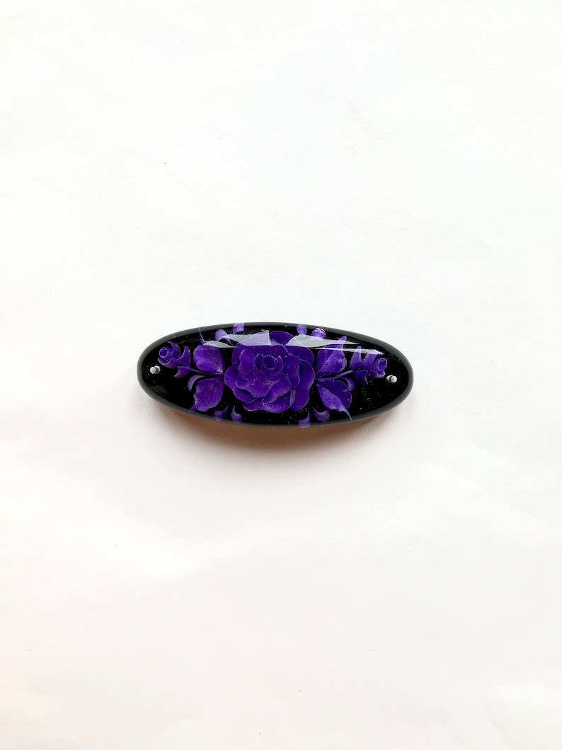 May include: A black oval hair clip with a purple floral design. The design features a large rose with smaller flowers and leaves.