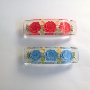 Könnte beinhalten: Zwei transparente Plastik-Haarklammern mit einer Reihe dreidimensionaler Rosen. Die obere Klammer hat rote Rosen und die untere Klammer hat blaue Rosen.