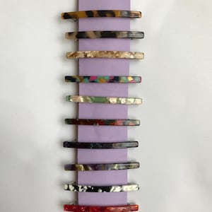 May include: A set of ten colorful hair clips with different patterns. The clips are made of a translucent material and have a curved shape. They are arranged vertically on a purple background.