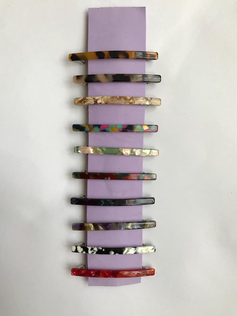 May include: A set of ten colorful hair clips with different patterns. The clips are made of a translucent material and have a curved shape. They are arranged vertically on a purple background.
