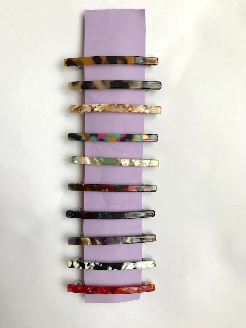 May include: A set of ten colorful hair clips. The clips are made of a translucent plastic material with different patterns and colors. The clips are arranged vertically on a purple background.