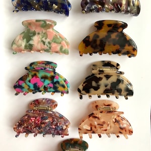 Pode incluir: Uma coleção de nove garras de cabelo com vários padrões de mármore e tartaruga em cores que incluem azul, verde, rosa, marrom, preto e bege. Cada garra tem um design articulado com dentes interligados e uma mola central de metal.