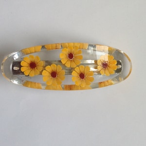 May include: A clear plastic hair clip with five yellow flowers with red centres. The clip is oval shaped.