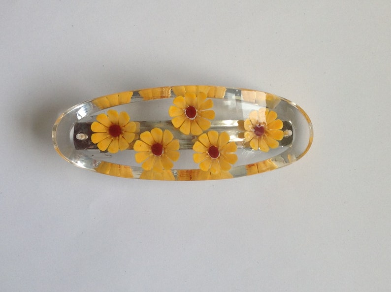 May include: A clear plastic hair clip with five yellow flowers with red centres. The clip is oval shaped.