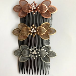 Op de afbeelding: Drie metalen haar kammen met bloemmotieven. De kammen zijn zwart, koper en zilver met strass in het midden van elke bloem.