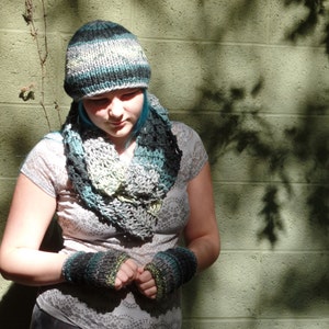 May include: A person wearing a gray and teal knitted hat and scarf with matching fingerless gloves. The hat and scarf are made with a chunky yarn and have a loose, open weave.