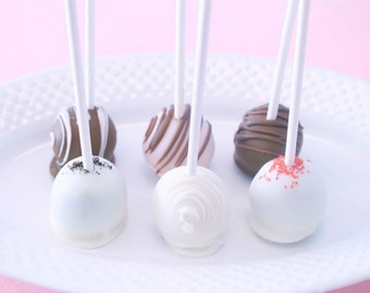 Puede incluir: Seis cake pops en un plato blanco. Los cake pops están cubiertos de chocolate blanco y decorados con chispas, chocolate y rayas blancas.