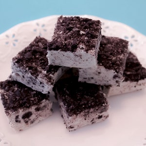 May include: A close-up of a stack of white chocolate fudge squares topped with crushed Oreo cookies. The fudge is on a white plate with a decorative lace pattern.