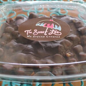 May include: A clear plastic container filled with chocolate covered nuts and candies. The container has a sticker on it that says "The Sweet Life, We Gotcha Covered" with a pink and blue umbrella graphic.