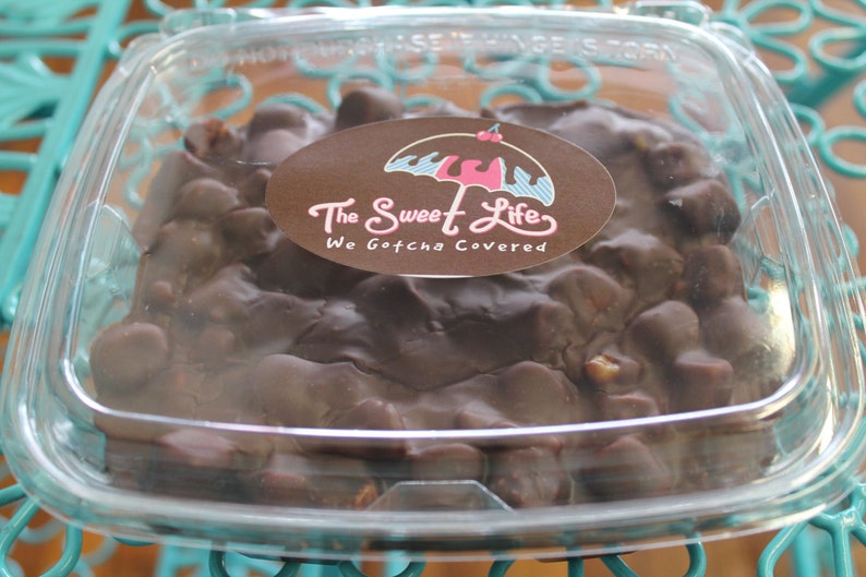 May include: A clear plastic container filled with chocolate covered nuts and candies. The container has a sticker on it that says "The Sweet Life, We Gotcha Covered" with a pink and blue umbrella graphic.