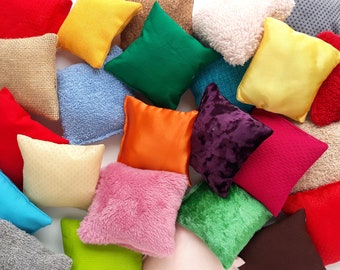 May include: A collection of small, square pillows in various colors and textures. The pillows include shades of red, yellow, green, orange, pink, and blue. Some pillows have smooth fabrics, while others are textured or plush.
