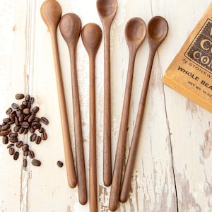 May include: A collection of six wooden spoons with long handles and rounded bowls. The spoons are a warm brown color and are arranged on a distressed white wooden surface. Coffee beans and a package of whole bean coffee are also present.