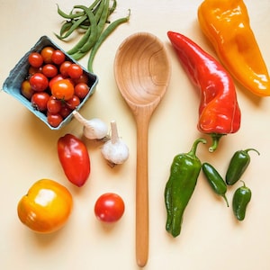 May include: A wooden spoon centered among fresh produce including a blue carton of cherry tomatoes, green beans, two garlic bulbs, a large yellow tomato, a red plum tomato, a red bell pepper, a yellow bell pepper, and several green chili peppers.