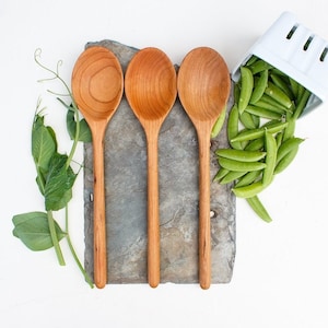 May include: Three wooden spoons with long handles and rounded bowls are arranged on a gray stone. Fresh green pea pods spill from a white container. Green leaves and tendrils are scattered around the spoons and stone, all set against a white background.