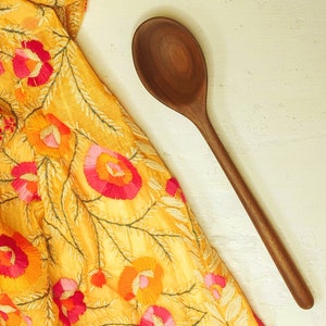 May include: A wooden spoon with a long handle rests on a white surface next to a yellow fabric with embroidered floral patterns in shades of pink, orange, and red. The spoon is made of dark wood.
