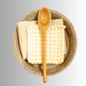 May include: A wooden spoon rests on a beige linen napkin and a yellow and white checkered potholder inside a rustic, round, brown bowl. The spoon is light brown and has a long handle. The potholder has a scalloped edge.