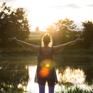 May include: A person stands with arms outstretched towards the setting sun over a lake. The person is wearing a patterned top and purple pants. The scene is set in a natural landscape with trees and fields, bathed in warm sunlight.