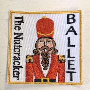 May include: A white embroidered patch with a gold border featuring a cartoon Nutcracker soldier in a red and gold uniform. The text "The Nutcracker" is on the left side of the patch and "BALLET" is on the right side.