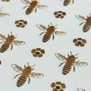 May include: Gold-colored bee and flower stickers on a white background. The bees have detailed wings and striped abdomens. The flowers are small and have a simple, five-petal design. These stickers are suitable for crafts and decorations.