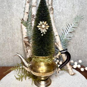 Puede incluir: Una tetera de latón vintage contiene un pequeño árbol de Navidad verde de cepillo para botellas, adornado con un broche de oro y cristal. La tetera tiene un asa negra y se asienta sobre una base decorativa. Decoración festiva navideña.