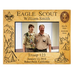 May include: A light-colored wooden picture frame featuring laser-engraved designs including mountains, tents, a campfire, and the Eagle Scout logo. The text reads Eagle Scout, William Smith, Once An Eagle Always An Eagle, Troop 123, January 13, 2018, Bakersfield, California.