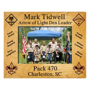 May include: A wooden framed photo featuring a group of Boy Scouts and leaders under a blue canopy. The frame includes text: "Mark Tidwell, Arrow of Light Den Leader," and "Pack 470, Charleston, SC." Names of scouts are listed on the frame.