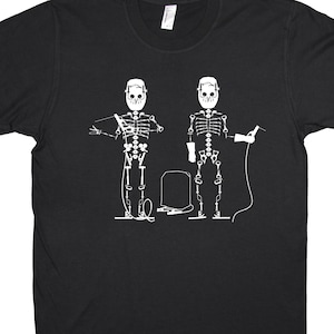 May include: Black t-shirt with a white graphic of two skeletons wearing welding masks and holding welding tools.