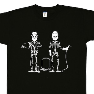 May include: A black short-sleeve t-shirt featuring a white graphic print of two skeletons wearing welding helmets. The skeletons are depicted with welding equipment, including a torch and cables, positioned on either side of a welding tank.