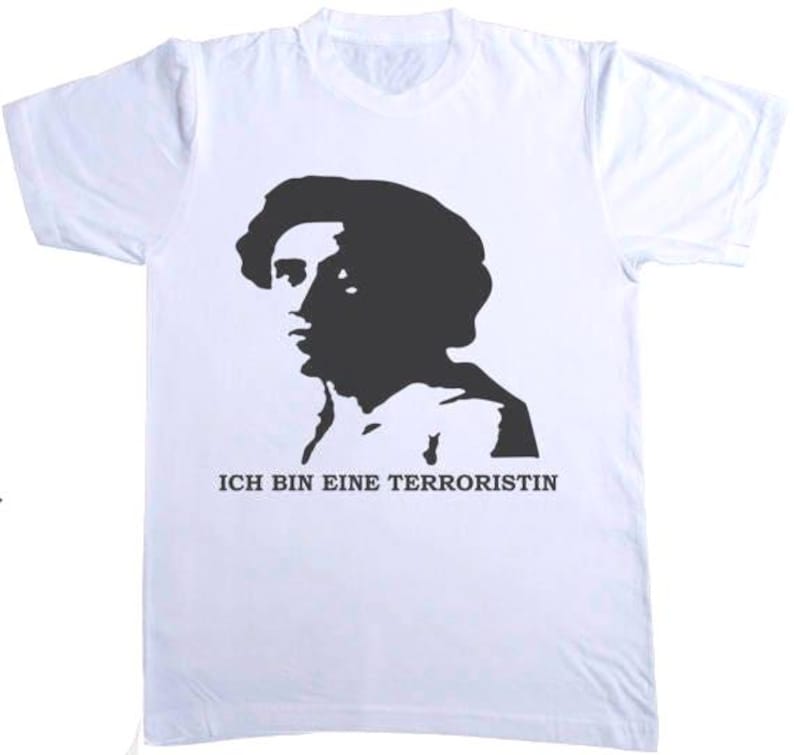 Peut inclure: T-shirt blanc avec une silhouette noire d'un profil de personne. Le texte allemand "ICH BIN EINE TERRORISTIN" est imprim&eacute; en noir sous l'image. Le t-shirt a des manches courtes et un col rond.