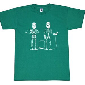 May include: A short-sleeve emerald green t-shirt featuring a white graphic print of two skeletons wearing welding helmets and gloves. The skeletons are depicted holding welding equipment with a central welding tank positioned between them.
