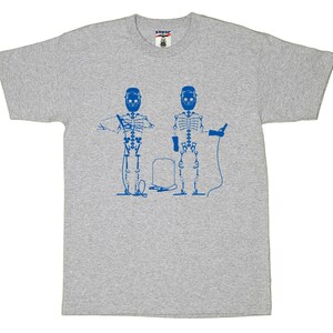 May include: A heather gray short-sleeve t-shirt featuring a blue graphic print of two skeletons wearing welding helmets. One skeleton holds a welding torch with a cable, while the other holds a pair of pliers. A welding tank sits between them.