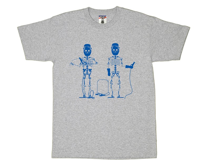 May include: A heather gray short-sleeve t-shirt featuring a blue graphic print of two skeletons wearing welding helmets. One skeleton holds a welding torch with a cable, while the other holds a pair of pliers. A welding tank sits between them.