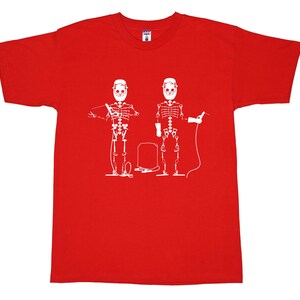 May include: A bright red short-sleeved t-shirt featuring a white graphic print of two skeletons wearing welding helmets. The skeletons are depicted holding welding equipment, with a welding machine positioned between them on the chest area.