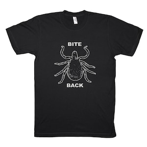 Puede incluir: Camiseta negra con un gráfico blanco de una garrapata y las palabras "BITE BACK" en negrita. El diseño está centrado en la parte delantera de la camiseta. La camiseta tiene cuello redondo y mangas cortas.