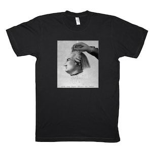 May include: Black t-shirt featuring a black and white graphic print. The graphic depicts a historical illustration of a severed head with a hand holding the hair. The text at the bottom of the graphic is blurry.