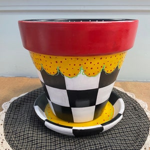 Puede incluir: Un macetero de terracota pintado en rojo, amarillo, negro y blanco con un patrón de tablero de ajedrez y un borde festoneado. El macetero está sentado sobre un posavasos de tela negro y blanco.