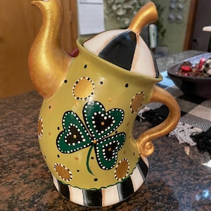 May include: A green ceramic teapot with a gold spout and handle. The teapot is decorated with a shamrock, gold dots, and black and white stripes. The lid is black and white with a gold knob.