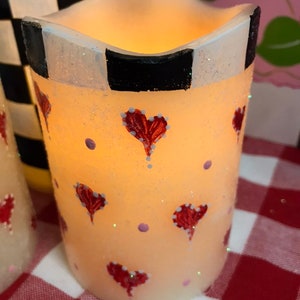 Puede incluir: Una vela blanca con un patrón de cuadros blanco y negro alrededor de la parte superior. La vela está decorada con corazones rojos y puntos rosas.