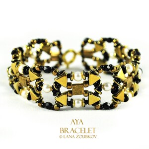 Puede incluir: Una pulsera dorada con cuentas negras y blancas y formas geométricas. La pulsera tiene un cierre y está adornada con pequeñas perlas. El texto "AYA BRACELET" es visible.